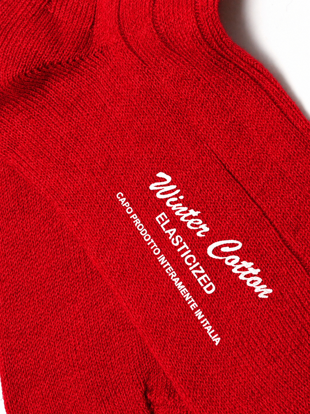 A close-up of a bright red knit fabric product, likely a sock or sleeve, with white printed text reading 'Winter Cotton' and 'ELASTICIZED' plus smaller Italian text. No person, animal, or real environment is visible, so this is Product/Packaging rather than Lifestyle. socks, cotton socks, high-knee socks, made in italy, handmade, artigianally made, fatto a mano, calzettoni invernali, winter cotton, calze di cotone, calze di cachemire, cachemire socks, cashmire socks