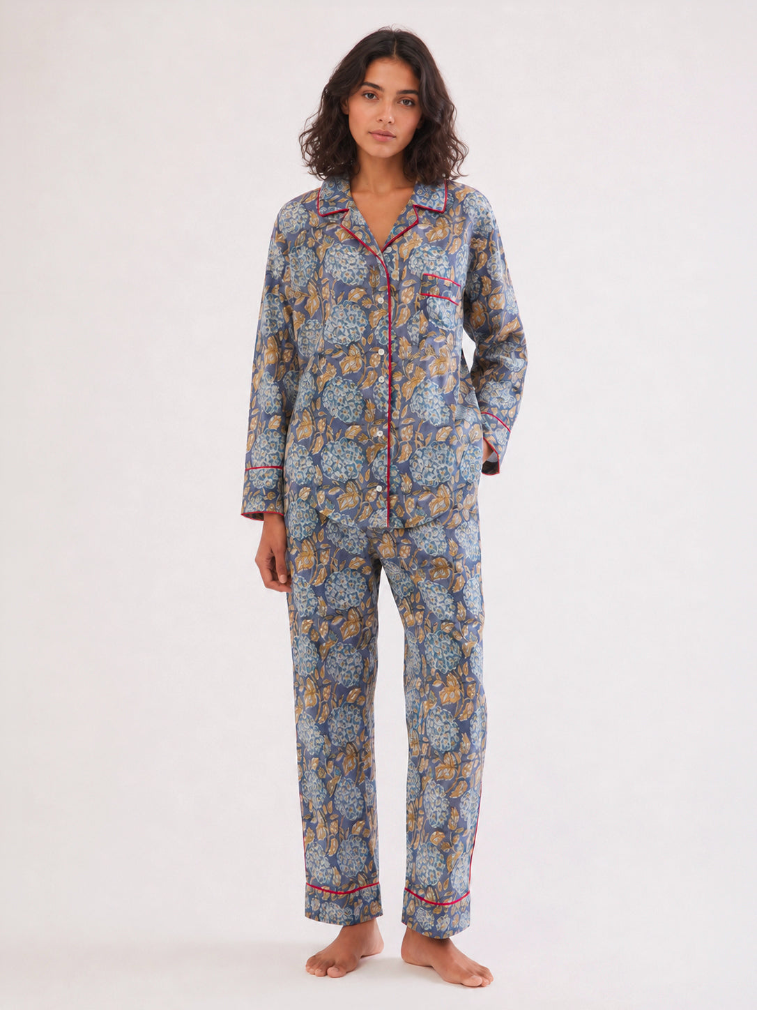 A woman modeling a two-piece pajama set with a blue, yellow, and orange floral pattern and red piping, standing against a plain white studio background. ETHICAL FASHION, CRAFTSMANSHIP, SUSTAINABLE FASHION, TRADITIONAL FASHION, ETHNIC FASHION, BOHEMIAN FASHION, MODA ETICA, MODA SOSTENIBILE, TECNICHE ARTIGIANALI, MODA TRADIZIONALE, MODA ETNICA, BOHEMIAN DRESS, MADE IN INDIA, HANDMADE IN INDIA, BLOCK PRINT, FLOWER PAJAMAS, PIGIAMA A FIORI, PIGIAMA DI COTONE, COTTON PAJAMA, SILK PAJAMA, PIGIAMA DI SETA, VESTAGL