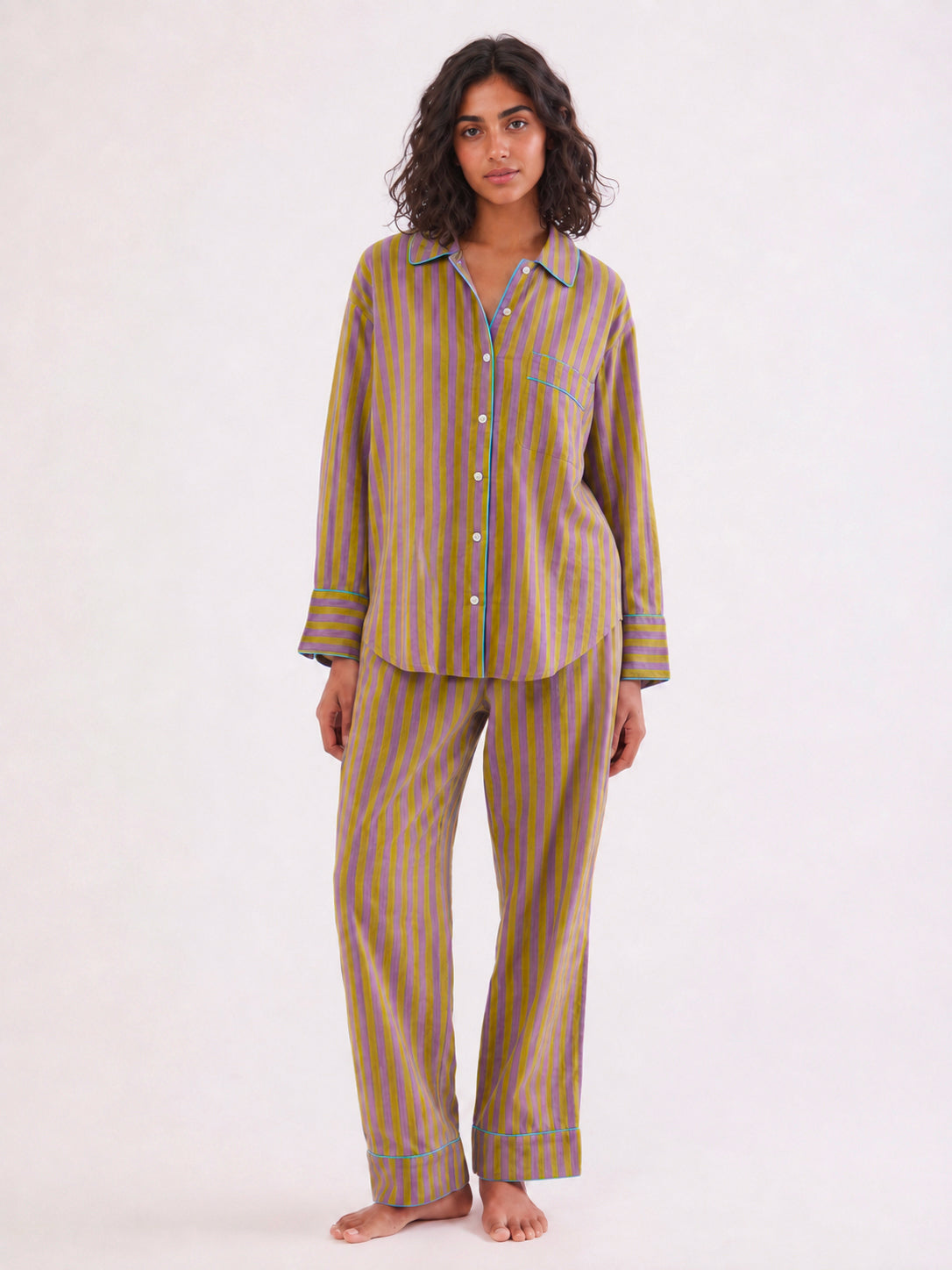 A woman stands barefoot against a plain white studio background wearing a matching pajama set with vertical stripes in muted olive green, mustard yellow, and blue, plus blue piping details. ETHICAL FASHION, CRAFTSMANSHIP, SUSTAINABLE FASHION, TRADITIONAL FASHION, ETHNIC FASHION, BOHEMIAN FASHION, MODA ETICA, MODA SOSTENIBILE, TECNICHE ARTIGIANALI, MODA TRADIZIONALE, MODA ETNICA, BOHEMIAN DRESS, MADE IN INDIA, HANDMADE IN INDIA, BLOCK PRINT, FLOWER PAJAMAS, PIGIAMA A FIORI, PIGIAMA DI COTONE, COTTON PAJAMA, 