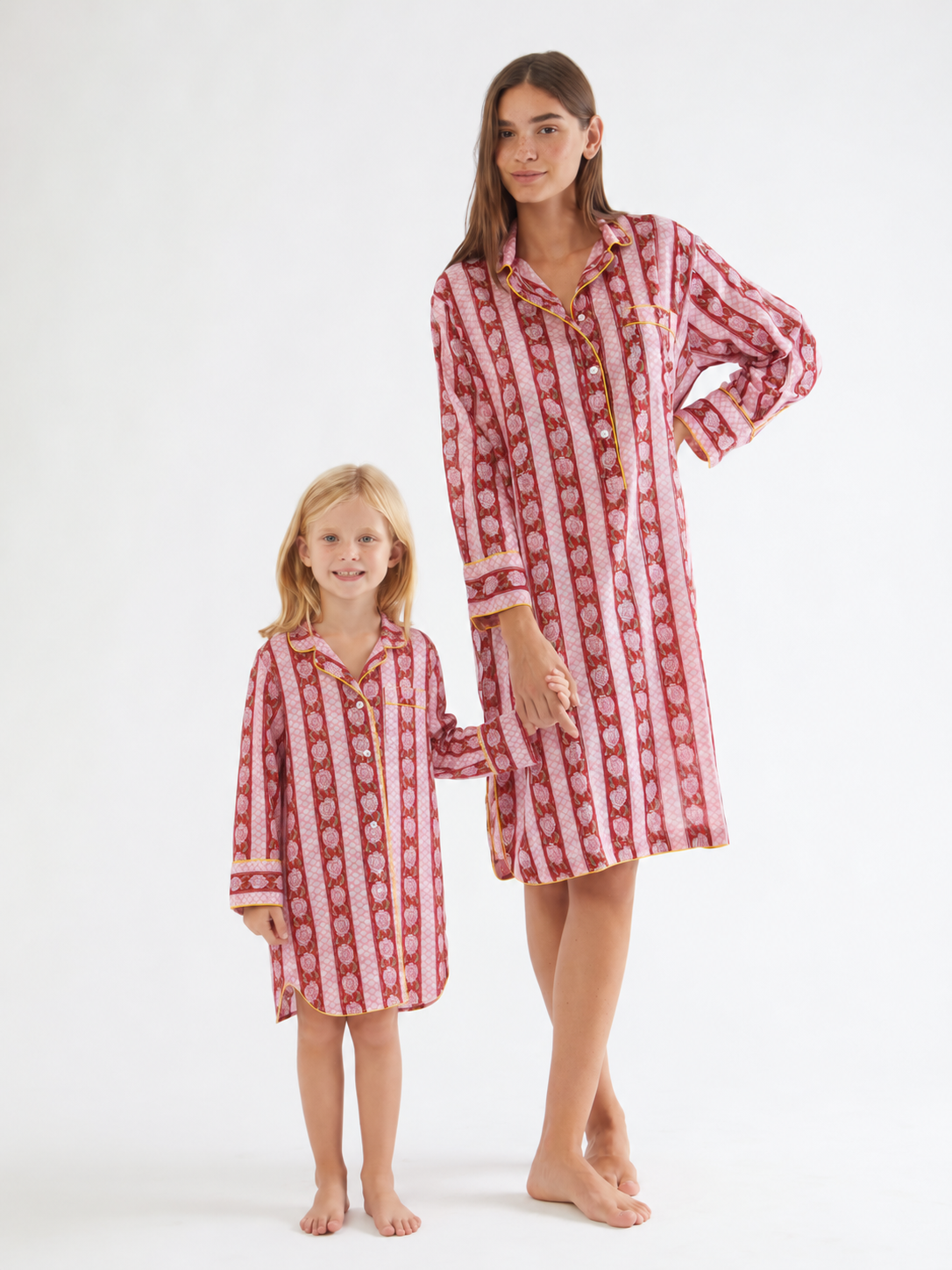 A woman and a young girl standing barefoot against a plain white studio background, both wearing matching long-sleeve button-front pajama dresses with vertical red and cream patterns. ETHICAL FASHION, CRAFTSMANSHIP, SUSTAINABLE FASHION, TRADITIONAL FASHION, ETHNIC FASHION, BOHEMIAN FASHION, MODA ETICA, MODA SOSTENIBILE, TECNICHE ARTIGIANALI, MODA TRADIZIONALE, MODA ETNICA, BOHEMIAN DRESS, MADE IN INDIA, HANDMADE IN INDIA, BLOCK PRINT, FLOWER PAJAMAS, PIGIAMA A FIORI, PIGIAMA DI COTONE, COTTON PAJAMA, SILK P
