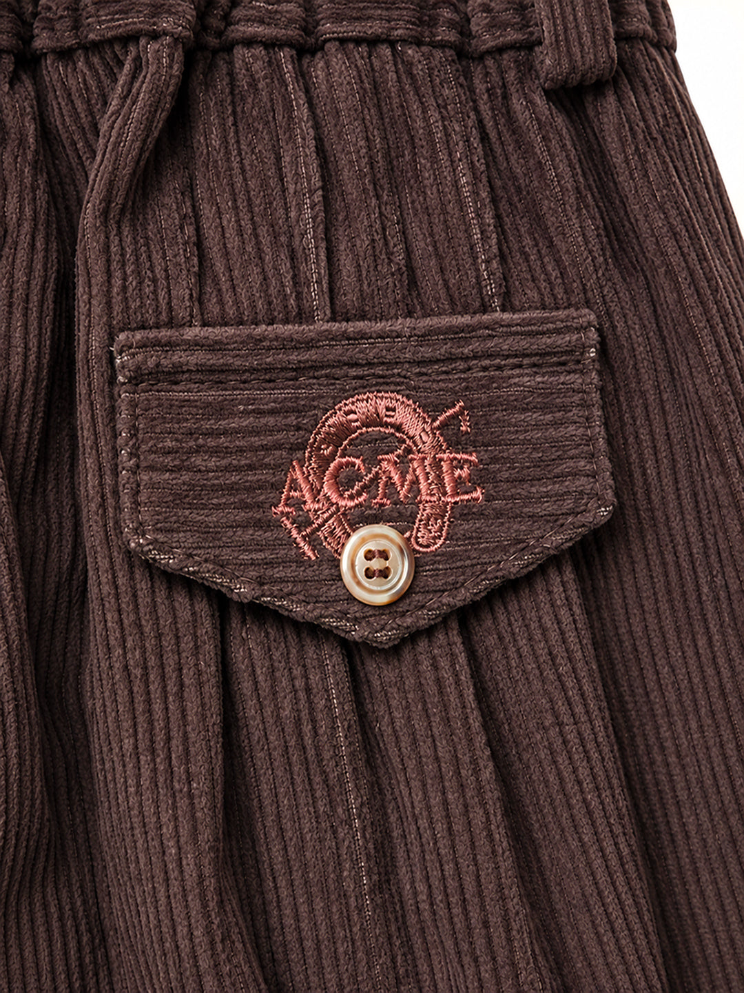 Close-up of a dark brown ribbed corduroy garment, likely a skirt or pants, featuring a front pocket with a button and an embroidered 'ACME' logo in pinkish-red thread. Argentine Bombacha Pants, horseback riding, traditional gaucho, Argentine cowboy attire. Soft and breathable fabrics, perfect pants for all-day wear. With a loose fit and elasticated waistband, they allow for plenty of movement, while the high waist provides extra coverage. Everyday look, perfect for both urban and countryside adventures. Cot