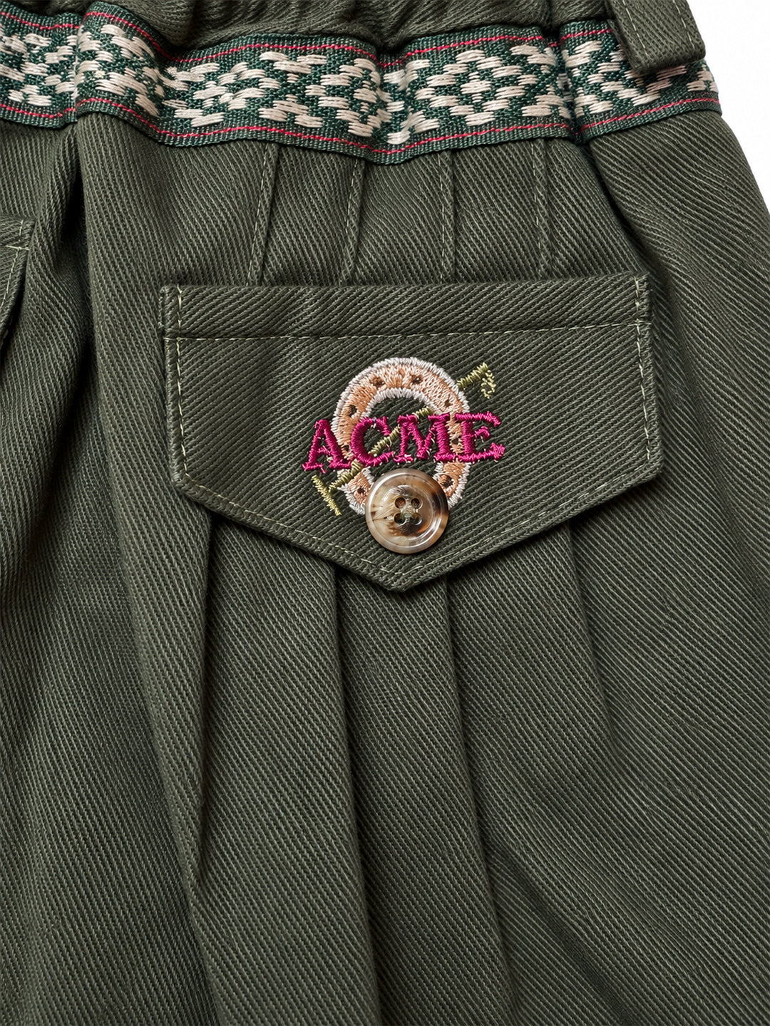 Close-up of olive green pleated fabric with a front pocket featuring a brown button and a circular embroidered patch with pink 'ACME' lettering. Argentine Bombacha Pants, horseback riding, traditional gaucho, Argentine cowboy attire. Soft and breathable fabrics, perfect pants for all-day wear. With a loose fit and elasticated waistband, they allow for plenty of movement, while the high waist provides extra coverage. Everyday look, perfect for both urban and countryside adventures. Cotton Corduroy, KIDS, PAN