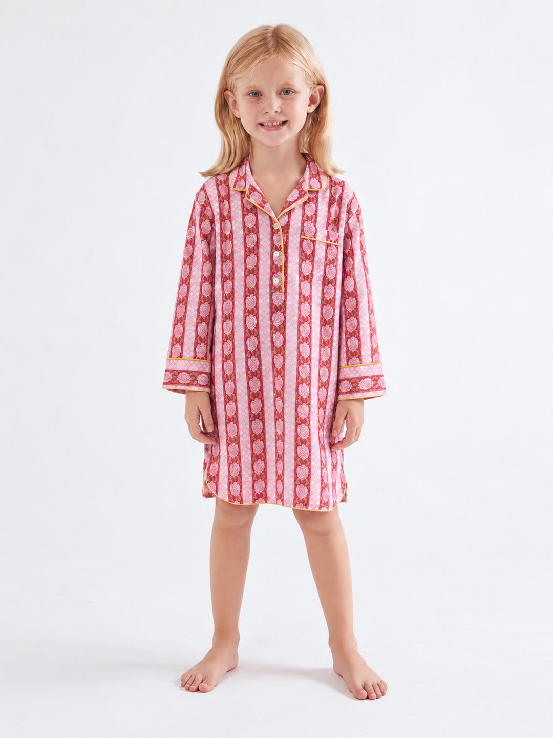 A young girl with blonde hair stands barefoot against a plain white studio background, wearing a pink and white vertically patterned pajama shirt with gold buttons. ETHICAL FASHION, CRAFTSMANSHIP, SUSTAINABLE FASHION, TRADITIONAL FASHION, ETHNIC FASHION, BOHEMIAN FASHION, MODA ETICA, MODA SOSTENIBILE, TECNICHE ARTIGIANALI, MODA TRADIZIONALE, MODA ETNICA, BOHEMIAN DRESS, MADE IN INDIA, HANDMADE IN INDIA, BLOCK PRINT, FLOWER PAJAMAS, PIGIAMA A FIORI, PIGIAMA DI COTONE, COTTON PAJAMA, SILK PAJAMA, PIGIAMA DI S