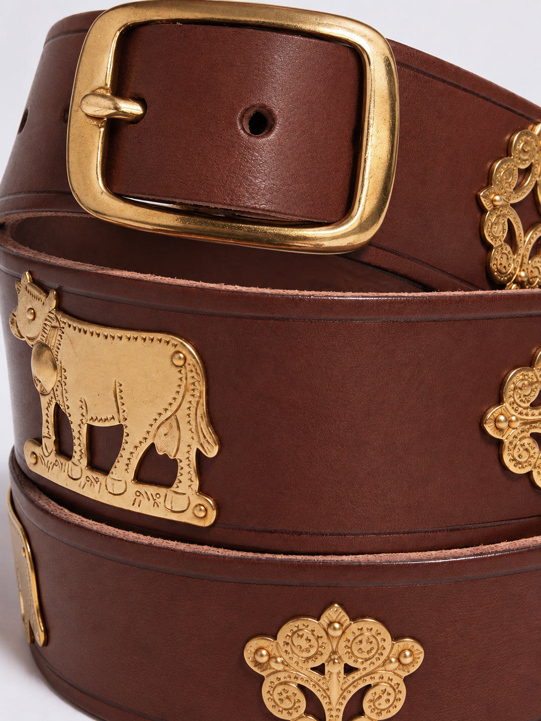 A close-up of a dark brown leather belt with ornate gold-tone metal hardware, including a large rectangular buckle, a cow motif, and decorative floral emblems. The Alpine Belt, Appenzeller belt, made in Switzerland, handmade, mountain belt, leather belt, cintura di pelle, cintura con le mucche, stelle alpine, cintura svizzera, cintura tirolese, cintura ampezzana, traditional belt, cow belt, kids belt, artisanal kids belt, artisanal belt, cinture da bambini, cinture artigianali.