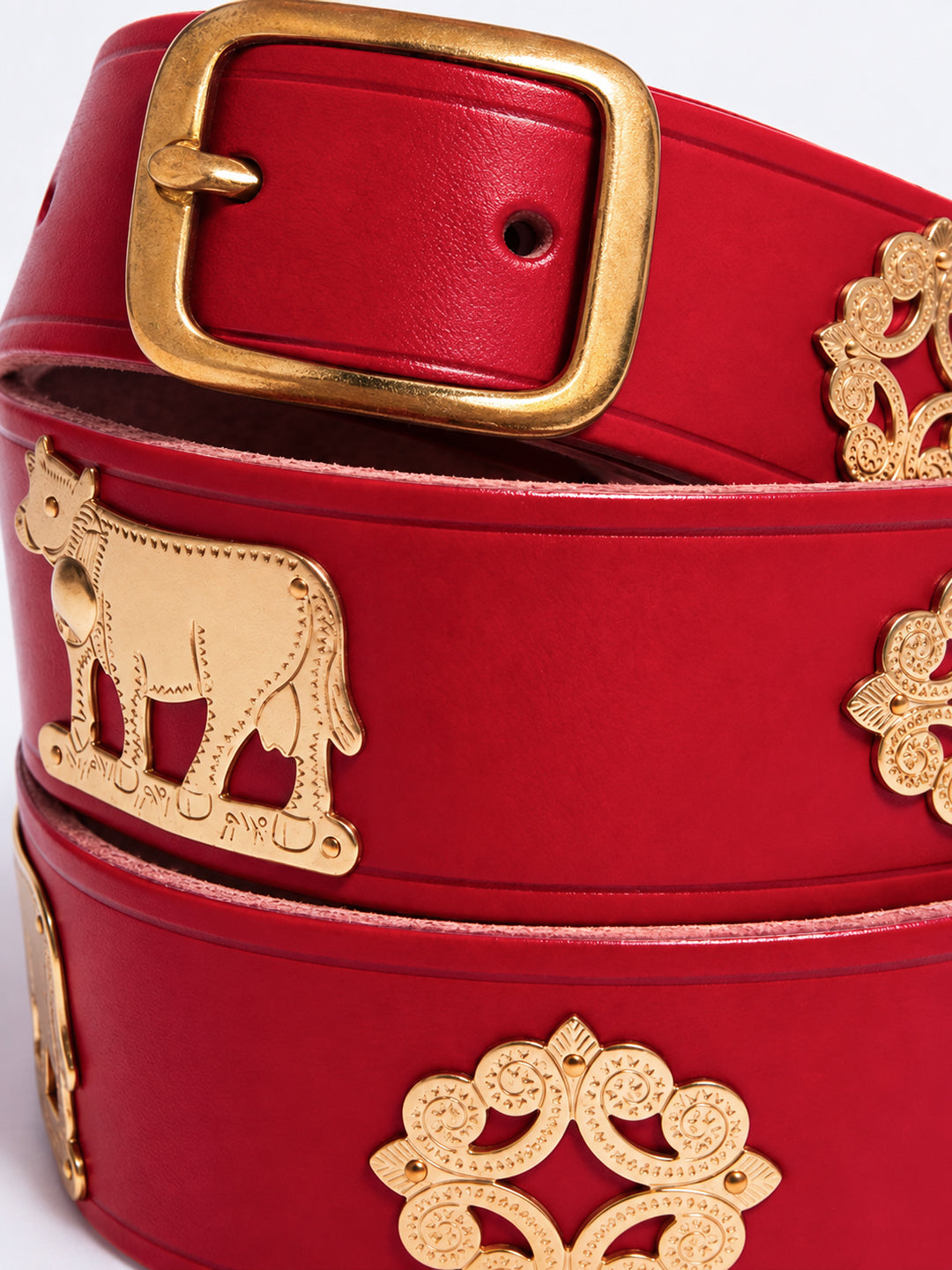 A close-up of a red leather belt featuring gold-tone metal accents, including a large square buckle, a cow-shaped emblem, and ornate filigree details. The Alpine Belt, Appenzeller belt, made in Switzerland, handmade, mountain belt, leather belt, cintura di pelle, cintura con le mucche, stelle alpine, cintura svizzera, cintura tirolese, cintura ampezzana, traditional belt, cow belt, kids belt, artisanal kids belt, artisanal belt, cinture da bambini, cinture artigianali.